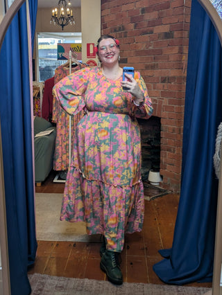Person smiling, taking a mirror selfie wearing a colorful floral dress in a room with a brick wall and blue curtains.