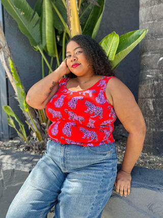 Woman wearing a red patterned top and blue jeans sitting outdoors with plants in the background