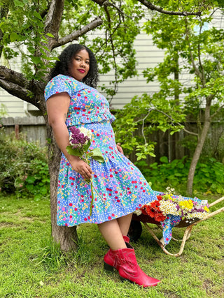 Woman in a colorful skirt and top holding flowers, standing next to a wheelbarrow with more flowers, in a garden setting.