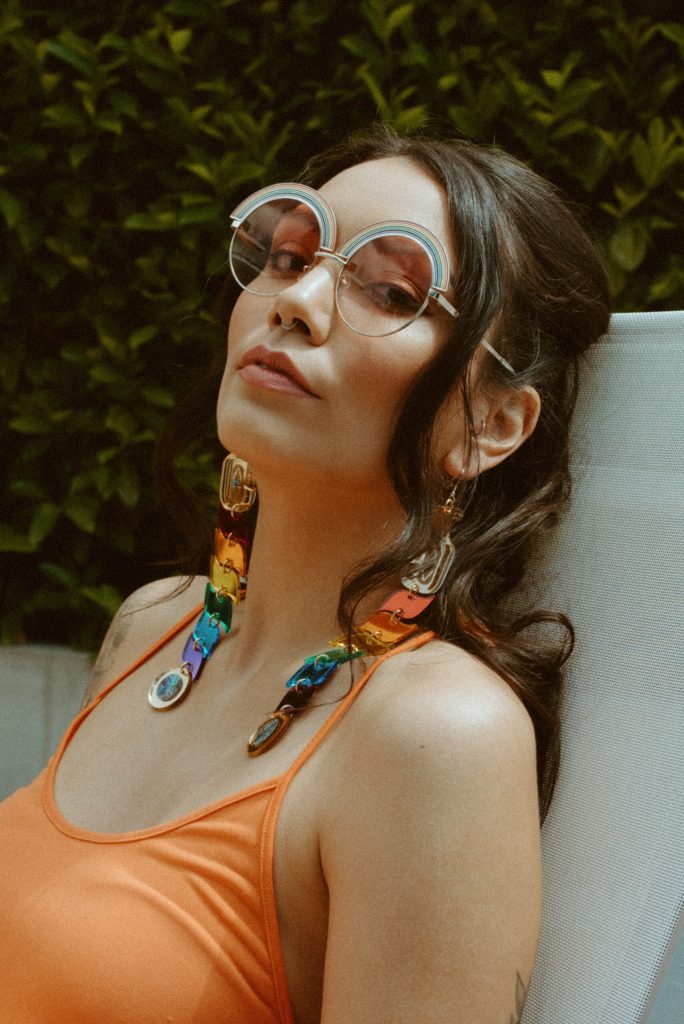 Person with long curled hair and big round sunglasses, reclining on a sun lounge, wearing long colorful dangly earrings cascading over their collarbones.