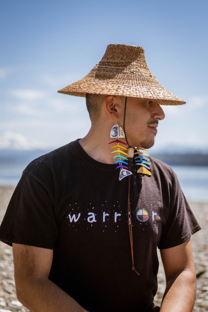 Person on the beach wearing large dangling earrings that resemble colorful fish skeletons. They also wear a woven grass hat and a black tshirt that says "warrior."