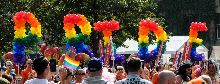 Rainbow-colored balloon sculptures that spell out PRIDE are held aloft, above a crowd of people waving various Pride and Equality flags.