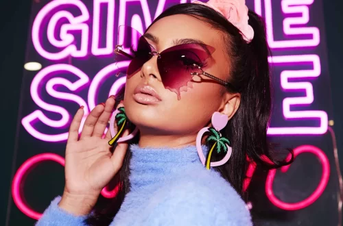 Portrait of a young femme person with pouty lips, medium skin tone and dark hair slicked back into a high ponytail with a pink scrunchie. She's wearing pink butterfly sunglasses and pink heart-shaped dangly earrings with a palm tree, and a lavender wool turtleneck. Behind her is a partly obscured pink neon sign that might say "Gimme some."