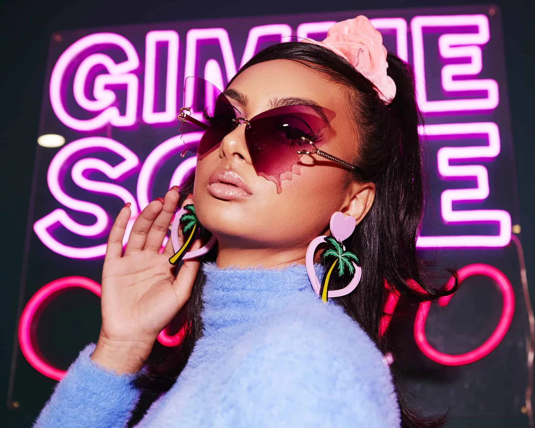 Portrait of a young femme person with pouty lips, medium skin tone and dark hair slicked back into a high ponytail with a pink scrunchie. She's wearing pink butterfly sunglasses and pink heart-shaped dangly earrings with a palm tree, and a lavender wool turtleneck. Behind her is a partly obscured pink neon sign that might say "Gimme some."