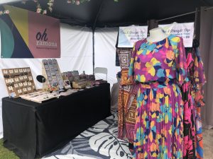 A market stall with colorful women's clothing on the right and a table of jewellery and paper goods on the left. Above the table hangs a banner reading "Oh Ramona"