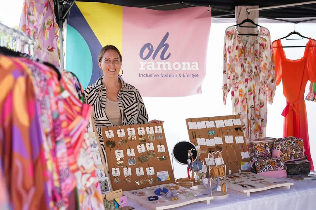 Cara stands smiling in her market stall stand, surrounded by colourful dresses, chunky jewellery and a promotional sign for "Oh Ramona – inclusive fashion & lifestyle."
