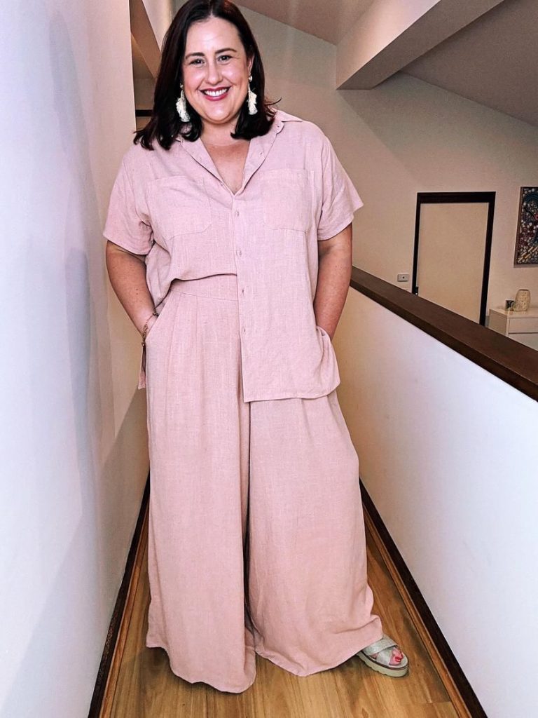 Amy smiles at the camera, looking very put together a matching pale pink set of a short-sleeved shirt and wide leg pants, with chunky earrings.