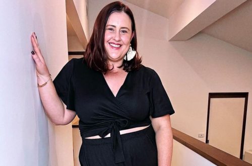 Stylist Amy smiles at the camera, looking put together and confident in a black wrap top and matching black pants, accessorised with a chunky earring.